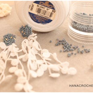 May include: A pair of handmade earrings with delicate white crocheted fringe and blue beaded clusters. The earrings are displayed on a wooden surface with a spool of white DMC Cordonnet Special thread, a small container of blue beads, and two earring backs.
