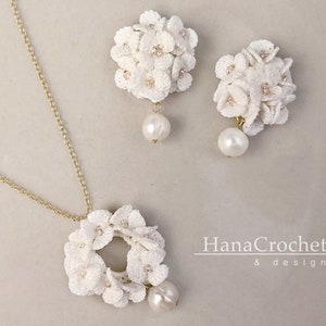 Peut inclure: Un ensemble de collier et de boucles d'oreilles en crochet blanc avec des accents de perles. Le collier pr&eacute;sente un pendentif circulaire avec un motif floral. Les boucles d'oreilles sont &eacute;galement florales et ont une perle qui pend de chacune. Le texte "HanaCrochet & design" est visible dans le coin inf&eacute;rieur droit de l'image.