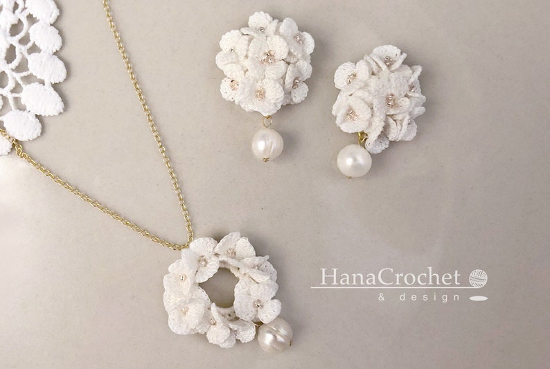 Peut inclure: Un ensemble de collier et de boucles d'oreilles en crochet blanc avec des accents de perles. Le collier pr&eacute;sente un pendentif circulaire avec un motif floral. Les boucles d'oreilles sont &eacute;galement florales et ont une perle qui pend de chacune. Le texte "HanaCrochet & design" est visible dans le coin inf&eacute;rieur droit de l'image.