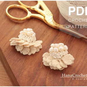 May include: Two delicate crocheted flower earrings with pearl accents. The earrings are on a wooden surface with a pair of gold scissors and a ball of yarn. The image includes the text "PDF CROCHET PATTERN" and "HanaCrochet & design".
