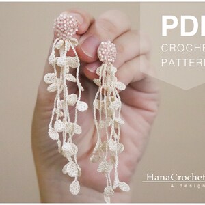 May include: PDF crochet pattern for a pair of long, delicate earrings with pink beads and white crocheted leaves. The earrings are held in a hand.