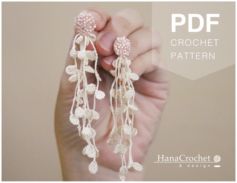 May include: PDF crochet pattern for a pair of long, delicate earrings with pink beads and white crocheted leaves. The earrings are held in a hand.
