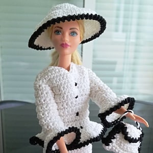 Può includere: Un completo da bambola bianco all'uncinetto con finiture nere, tra cui cappello, giacca e gonna. La bambola tiene anche una borsetta abbinata. Il completo è fatto a mano e presenta un punto strutturato.