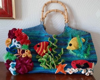 May include: A blue crocheted handbag with a bamboo handle, decorated with colorful crochet coral, fish, and seaweed. The bag features a rectangular shape and a striped pattern in shades of blue and green.