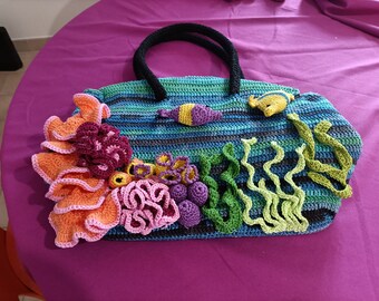 May include: Crocheted tote bag with a marine theme. The bag features a blue and green striped background, adorned with colorful coral, seaweed, and fish. The handles are black.