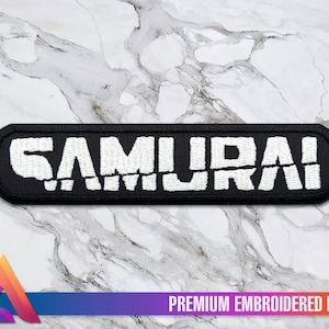Peut inclure: Écusson brodé noir avec le mot "SAMOURAÏ" en lettres blanches. L'écusson est rectangulaire et a un aspect légèrement vieilli. Le texte "PREMIUM EMBROIDERED PATCH" est imprimé sous l'écusson.