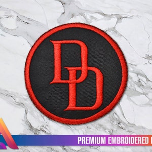 Peut inclure: Patch brodé rouge avec un fond noir et la lettre "D" dans une police stylisée. Le patch est circulaire et a une bordure rouge. Le texte "PREMIUM EMBROIDERED PATCH" est imprimé sous le patch.