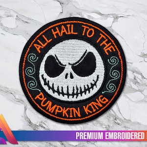 Könnte beinhalten: Ein schwarzer und weißer gestickter Aufnäher mit einem runden Design. Die Mitte zeigt ein weißes Jack Skellington Gesicht auf schwarzem Hintergrund. Der Text um das Gesicht lautet "All Hail To The Pumpkin King" in orangem und weißem Garn.