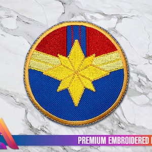 Peut inclure: Un patch rond brodé représentant le logo de Captain Marvel. Le logo est une étoile jaune sur fond rouge et bleu avec une bordure dorée. Le texte "PREMIUM EMBROIDERED PATCH" est sous le patch.