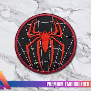 Könnte beinhalten: Roter gestickter Spinnennetz-Patch mit einer roten Spinne in der Mitte. Der Patch hat einen burgunderfarbenen Rand und befindet sich auf einem weißen Marmoruntergrund. "PREMIUM EMBROIDERED PATCH" steht in weißer Schrift auf schwarzem Hintergrund am unteren Rand des Bildes.