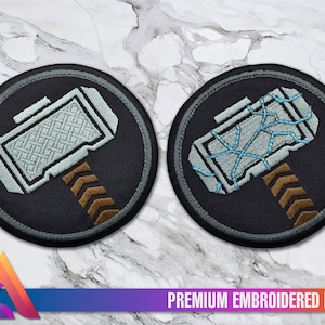 Peut inclure: Deux patchs brodés représentant une image stylisée du marteau de Thor, Mjolnir. Un patch a un marteau gris et blanc avec un manche brun, tandis que l'autre a un marteau gris et bleu avec un manche brun. Les deux patchs ont une bordure circulaire grise.