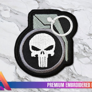 Può includere: Un patch ricamato in bianco e nero con un teschio e un design a forma di granata. Il teschio è bianco con occhi neri e un contorno nero. La granata è nera con un anello argentato e una miccia grigia. Il patch è etichettato "PREMIUM EMBROIDERED PATCH".