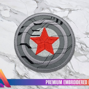 Könnte beinhalten: Ein grauer gestickter Aufnäher mit einem roten Stern in der Mitte. Der Stern ist von einem weißen Kreis umgeben, der wiederum von einem grauen Kreis umgeben ist. Der äußere Kreis hat ein graues und schwarzes Design, das einer Leiterplatte ähnelt. Der Aufnäher trägt unten den Schriftzug "PREMIUM EMBROIDERED PATCH".