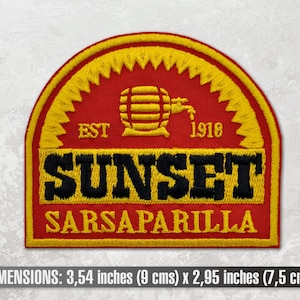 Op de afbeelding: Een geborduurde patch in rood, geel en zwart met de tekst "EST 1918 SUNSET SARSAPARILLA". De patch heeft een gestileerde afbeelding van een vat met een kraan. De patch is 9 cm bij 7,5 cm.