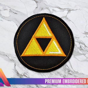 May include: A round, black embroidered patch featuring a yellow and white Triforce symbol. The patch has a yellow border and the text "PREMIUM EMBROIDERED PATCH" at the bottom. The patch is on a white and gray marble surface.