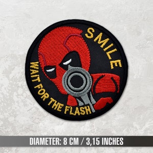 May include: A black and red embroidered patch with a masked figure holding a gun. The text "WAIT FOR THE FLASH" and "SMILE" are written around the figure. The patch is 8 cm / 3.15 inches in diameter.