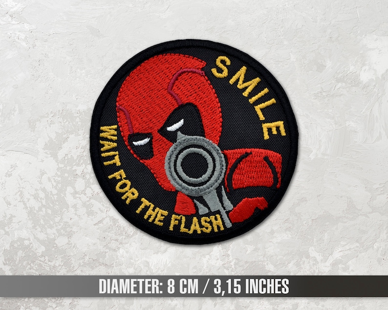 May include: A black and red embroidered patch with a masked figure holding a gun. The text "WAIT FOR THE FLASH" and "SMILE" are written around the figure. The patch is 8 cm / 3.15 inches in diameter.