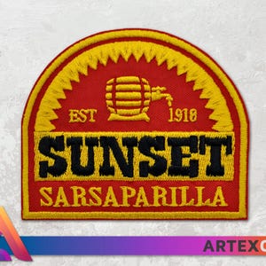 Op de afbeelding: Een geborduurde patch in rood, geel en zwart met de tekst "EST. 1918 SUNSET SARSAPARILLA" en een gestileerd beeld van een vat met een kraan.
