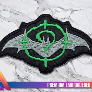 Könnte beinhalten: Grün-grauer gestickter Aufnäher mit einer Fledermaus-Silhouette in einem Ziel mit einem Fragezeichen in der Mitte. Der Text "PREMIUM EMBROIDERED PATCH" befindet sich unter dem Aufnäher.