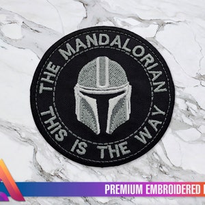 Peut inclure: Un patch brodé noir et gris avec un casque mandalorien au centre. Le texte autour du casque est "The Mandalorian This is the Way".