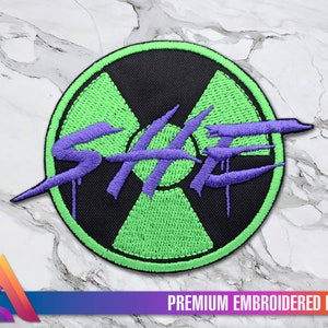 Può includere: Una toppa ricamata verde e nera con un logo "SHE" viola all'interno di un simbolo di radiazione stilizzato. La toppa è etichettata "PREMIUM EMBROIDERED PATCH".