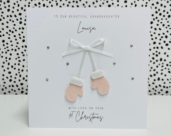 Op de afbeelding: Een witte kaart met roze glitter wanten en een wit lint. De kaart zegt "To our beautiful granddaughter Louisa with love on your 1st Christmas".