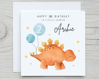 May include: A white card with a dinosaur illustration and the text "Happy 2nd Birthday to a special grandson Archie". The dinosaur is orange with yellow spikes and is holding two blue balloons. There are yellow stars scattered around the dinosaur.