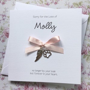 May include: A white sympathy card featuring the text Sorry for the Loss of Molly, no longer by your side but forever in your heart. A pale pink satin ribbon bow is attached to the center, adorned with a silver-toned angel wing and paw print charm.