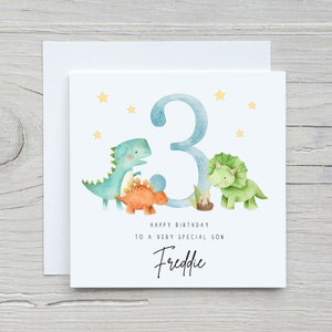 May include: A white card with a blue number 3 and three dinosaurs. The dinosaurs are green, orange, and blue. The card says "Happy Birthday to a very special son Freddie".