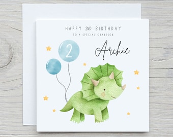 May include: A white card with a green dinosaur holding two blue balloons. The card says "Happy 2nd Birthday to a special grandson Archie".