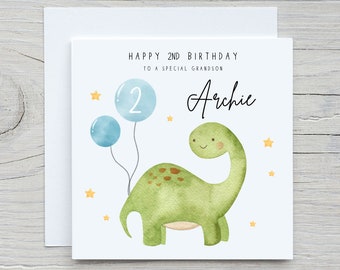 May include: A white card with a green dinosaur holding two blue balloons. The card says "Happy 2nd Birthday to a special grandson Archie".