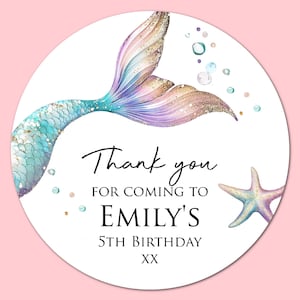 May include: A circular sticker featuring a watercolour mermaid tail in shades of teal, purple, and gold glitter. The text reads: Thank you FOR COMING TO EMILY'S 5TH BIRTHDAY XX. A small starfish is positioned on the right side with scattered bubbles.
