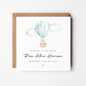 May include: A square greeting card featuring a watercolor illustration of a light blue hot air balloon with a small bear in the basket, flanked by clouds. Text reads: WELCOME TO THE WORLD, River Arthur Harrison, WEDNESDAY 12TH MAY 2025, with a blue heart.