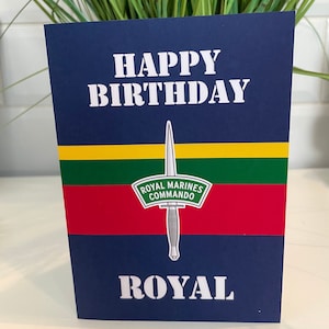 May include: A navy blue birthday card with white text reading "HAPPY BIRTHDAY" and "ROYAL". The card features a dagger emblem and the text "ROYAL MARINES COMMANDO" above red, green, and yellow stripes.