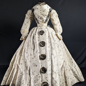 May include: A white and brown striped dress with a floral pattern. The dress has a black lace trim and a black ribbon around the waist. There are five black and gold buttons down the back of the dress.