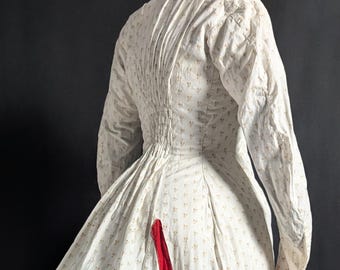 May include: A long-sleeved white dress featuring a small repeating floral pattern in light brown. The garment includes a gathered back bustle, a high neckline, and prominent horizontal red fabric bands along the skirt and hemline.