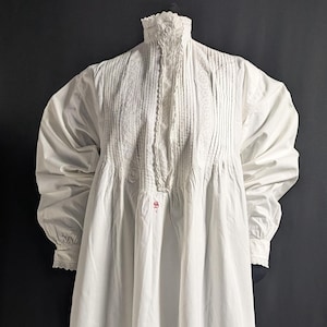 May include: A long, white nightgown with a high, lace-trimmed collar and long sleeves. The bodice features vertical pleats and delicate lace detailing. The sleeves end in lace cuffs. The nightgown is made of a soft, flowing fabric.
