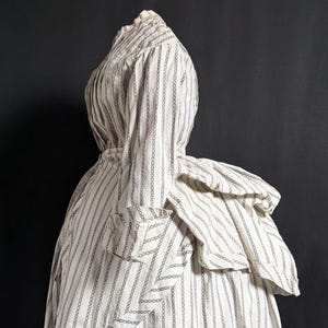 Könnte beinhalten: Ein weißes Vintage-Kleid mit vertikalem, schwarzem Kettengliedmuster. Das Kleid hat einen hohen Ausschnitt, lange Ärmel und ein geschichtetes, gerüschtes Detail am Rücken. Der Rock ist weit und reicht bis zu den Knöcheln.