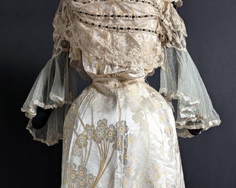 Puede incluir: Un vestido antiguo de color marfil con bordado floral. El vestido presenta un cuello alto con detalles de encaje, mangas transparentes y una falda amplia. La falda tiene un diseño floral y un lazo decorativo en el dobladillo.