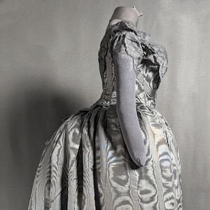 May include: A vintage-style, floor-length gown in shades of gray, featuring a unique, swirling pattern. The dress has a fitted bodice with lace detailing and short sleeves. The skirt is full and flowing, creating a dramatic silhouette. The fabric has a subtle sheen.