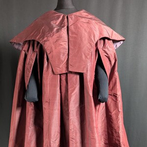 May include: A burgundy-colored costume cape with a structured collar and long, flowing fabric. The cape has a shiny, textured appearance and is displayed on a mannequin. Black sleeves are visible beneath the cape.