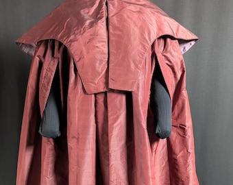 May include: A burgundy-colored costume cape with a structured collar and long, flowing fabric. The cape has a shiny, textured appearance and is displayed on a mannequin. Black sleeves are visible beneath the cape.