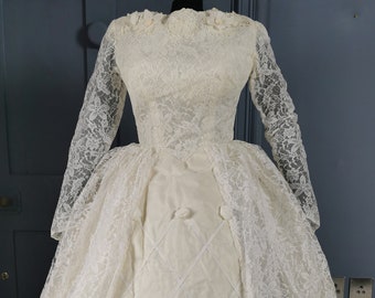 May include: A vintage white lace wedding dress with long sleeves and a full skirt. The dress has a fitted bodice and a gathered skirt. The skirt is lined with a white fabric and has a diamond pattern.