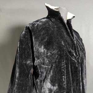 May include: A dark grey velvet jacket with a high collar and flared sleeves. The jacket has a soft, textured appearance and a contrasting white lining at the collar. The garment is displayed on a mannequin, showcasing its shape and design.