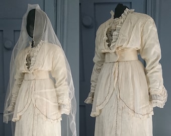 Puede incluir: Un vestido de novia blanco vintage con un corpiño de encaje y una falda larga. El vestido tiene un corpiño ajustado con una superposición de encaje y una falda larga y fluida con cola. El vestido está adornado con delicados bordados y una faja de satén.
