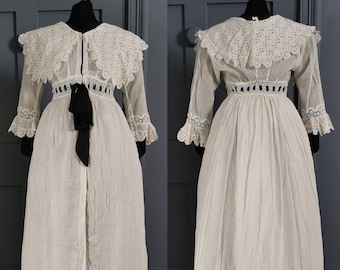 May include: A white vintage dress with a high, lace-trimmed collar and long sleeves. The dress features a detailed lace bodice, a black ribbon belt, and a long, flowing skirt. The dress is displayed on a black mannequin.