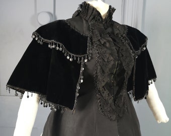May include: A black velvet cape with a lace trim and beaded fringe. The cape is worn over a black dress with a high collar.