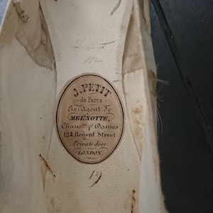 May include: Close-up of a white shoe insole with a brown label that reads "J. PETIT de Paris Ex-Agent de MELINOTTE, Chauss. p. Dames. 124, Regent Street Private door LONDON."