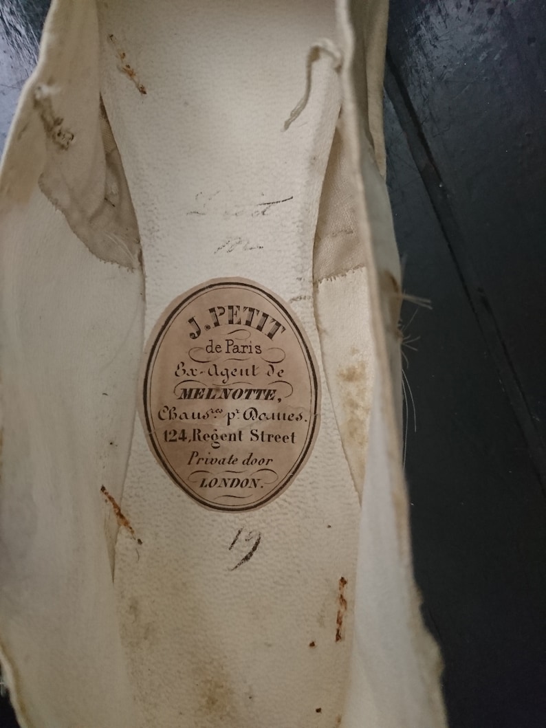 May include: Close-up of a white shoe insole with a brown label that reads "J. PETIT de Paris Ex-Agent de MELINOTTE, Chauss. p. Dames. 124, Regent Street Private door LONDON."