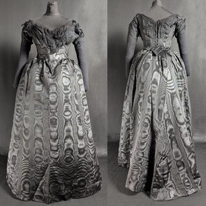 May include: A vintage, floor-length gown displayed on a mannequin. The dress features a patterned skirt and a detailed bodice with short sleeves. The fabric has a unique, swirling design in shades of gray and silver, creating a textured appearance. The dress is a historical garment.